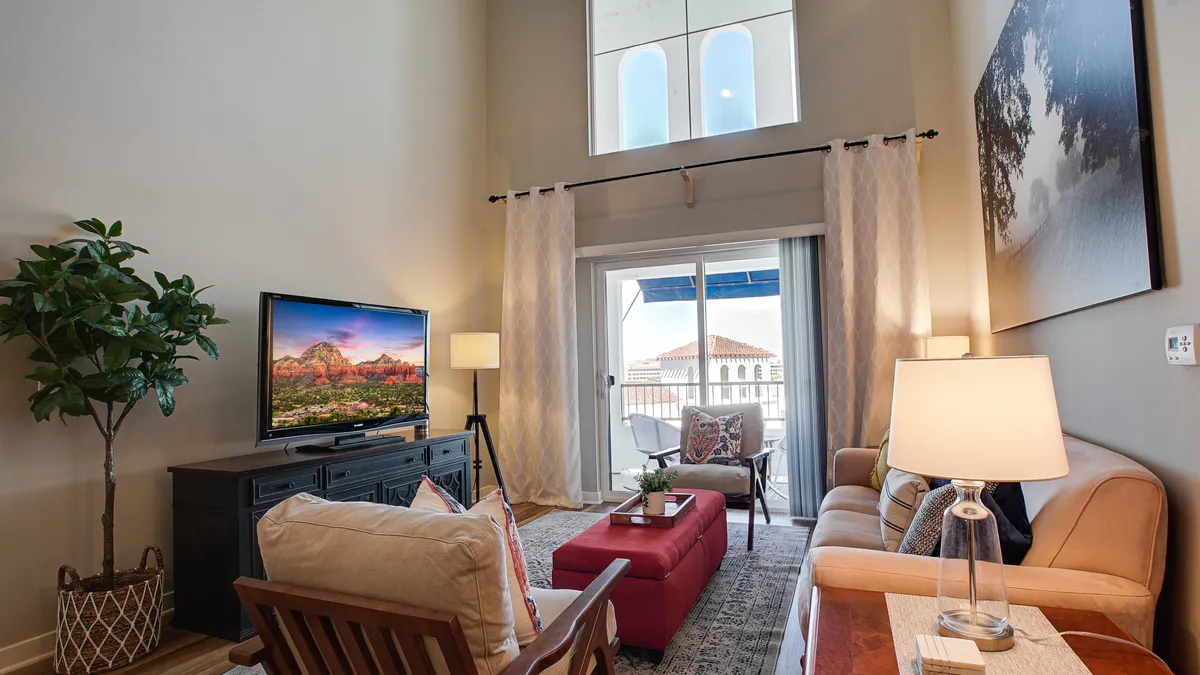 A bright, comfortable living area featuring a plush sofa, coffee table, and television, with large curtains framing a view onto the private balcony. The atmosphere is inviting, perfect for relaxing evenings or socializing, while the balcony hints at easy indoor-outdoor living.
