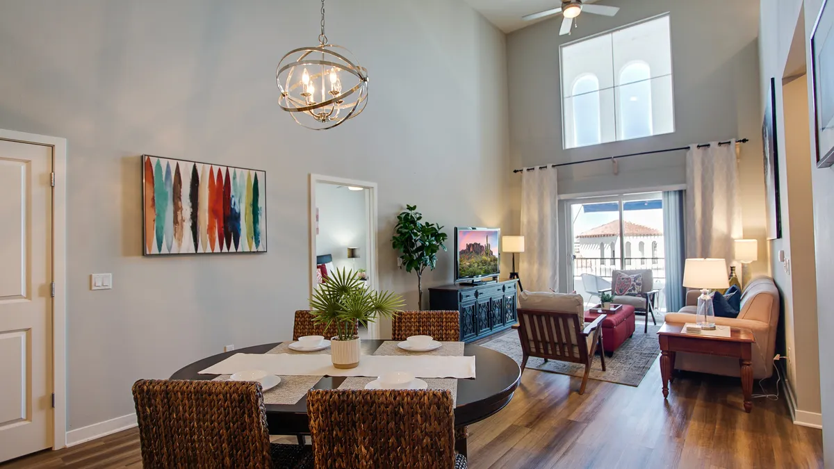 This interior image showcases a bright, open-concept living and dining area featuring a dining table set for six beneath a modern chandelier, a comfortable sofa, large windows that bring in natural light, and decorative artwork. The windows offer glimpses of the balcony and scenic views beyond, reinforcing the visual connection to the outdoors.