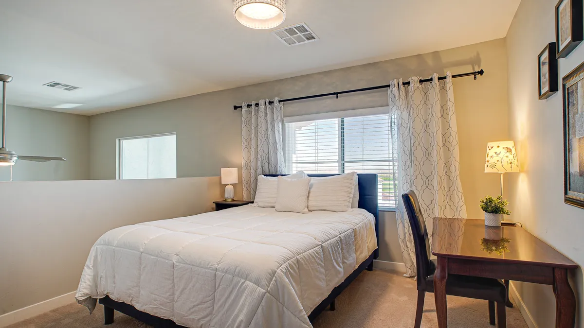 A bright, versatile bedroom features a cozy bed, a desk positioned near the window for natural light, and a serene atmosphere. The multi-purpose design suggests suitability for sleeping, working, or quiet retreat.