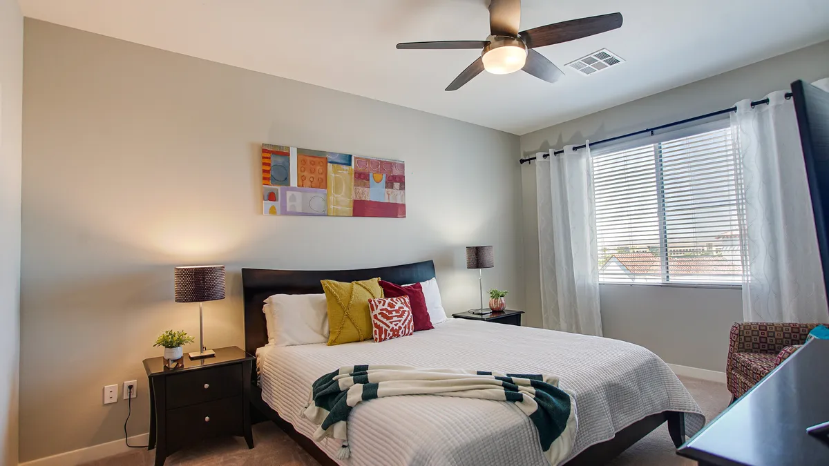 A serene bedroom with a comfortable bed, soft linens, art above the headboard, and sunlight streaming in through a window. Side tables and cozy accents create a restful atmosphere, ideal for relaxation and privacy.