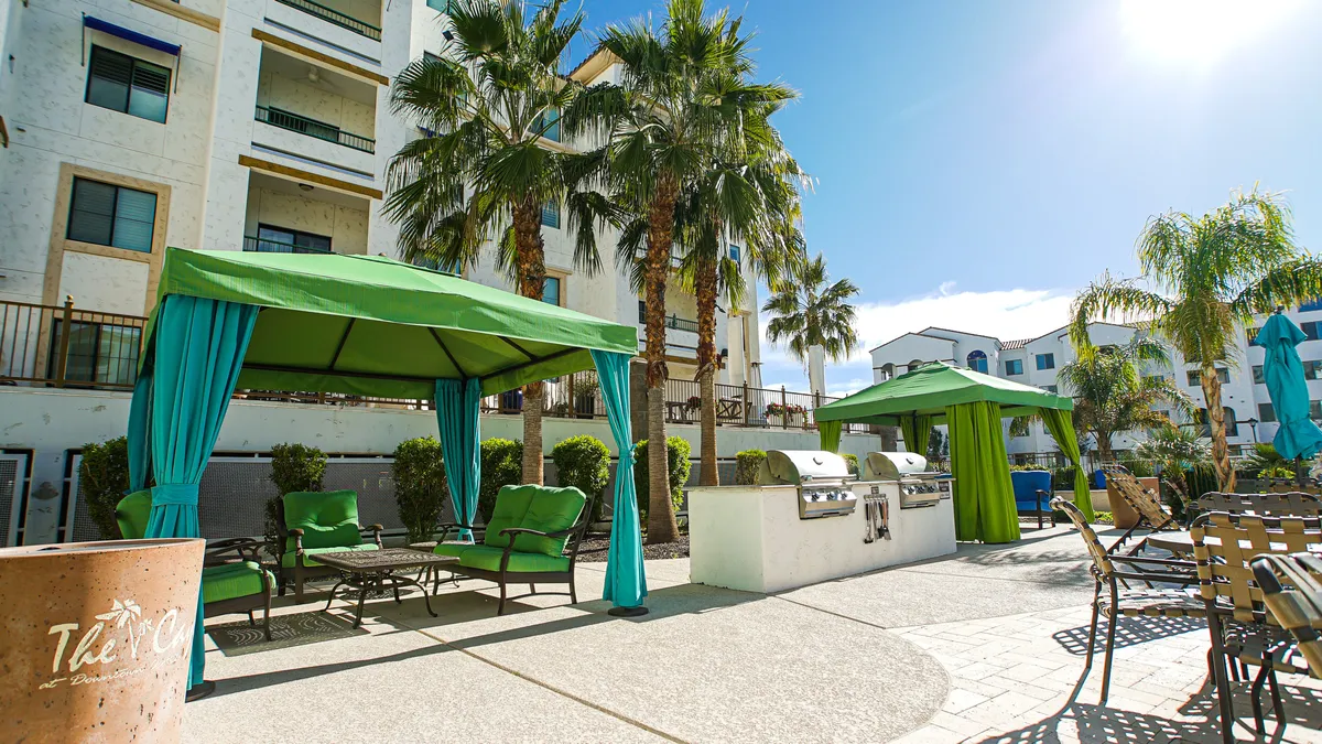 An outdoor communal area with green canopies, lounge chairs, a grill area, and palm trees. The space is designed for barbecuing, sunbathing, and socializing in a lush, resort-like environment.