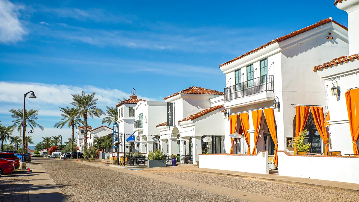 A cheerful outdoor setting with colorful orange curtains, palm trees, modern white buildings, and multiple seating areas. People can be seen enjoying meals outdoors and socializing, suggesting a lively community with opportunities for dining and gathering.