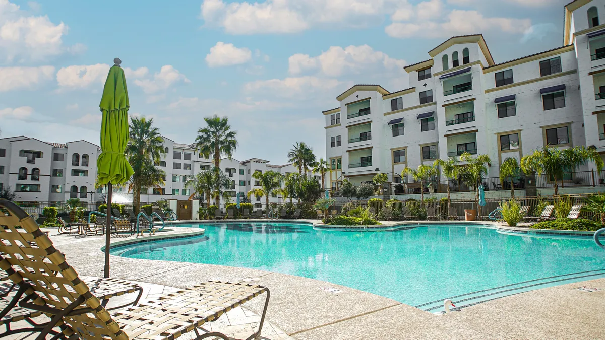 A sparkling swimming pool surrounded by palm trees, poolside loungers, and green umbrellas, with apartment buildings visible in the background. The atmosphere is inviting, with ample space for relaxation, sunbathing, and socializing outdoors.