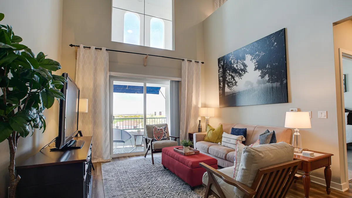 A cozy living room is pictured here, anchored by a plush sofa and a vibrant red ottoman, complemented by large wall art and a television. The room opens to a balcony through a glass door, letting in light and providing a seamless transition to outdoor views of the mountains or pool.