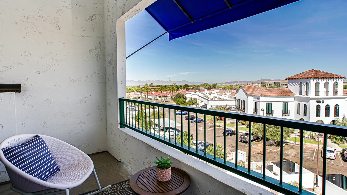 A private balcony scene featuring a round chair, small table with a plant, and serene views under a blue awning. The mood is tranquil and inviting, suggesting a comfortable spot to relax and enjoy the outdoors.