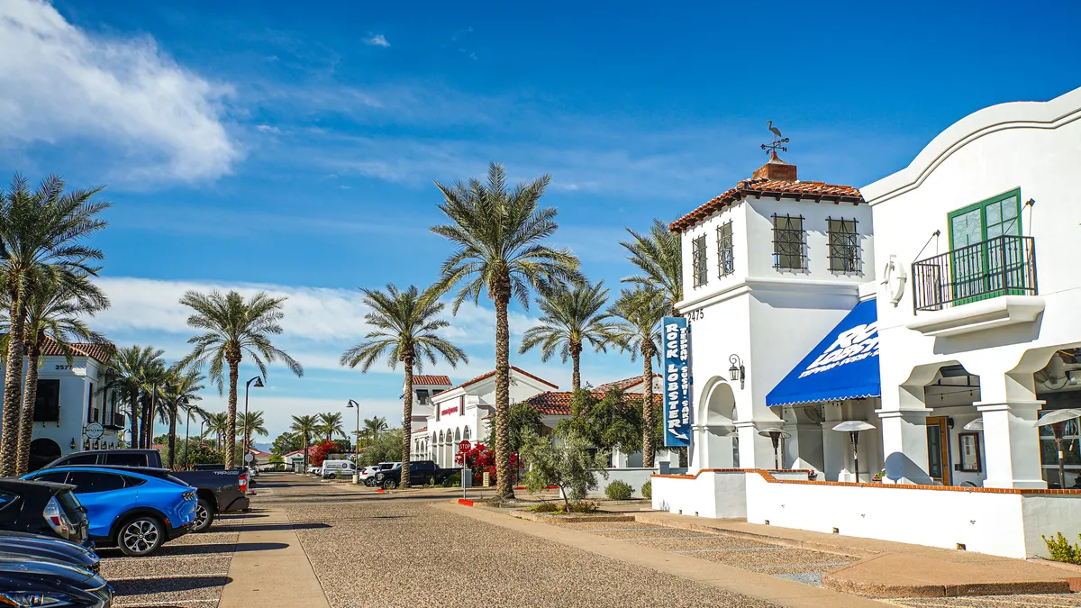 A lively exterior scene featuring palm trees, vibrant buildings with blue awnings, and a bustling sidewalk with people walking and cars parked nearby. The atmosphere evokes a sense of urban energy and accessibility, with clear references to shopping, dining, and enjoying local amenities.
