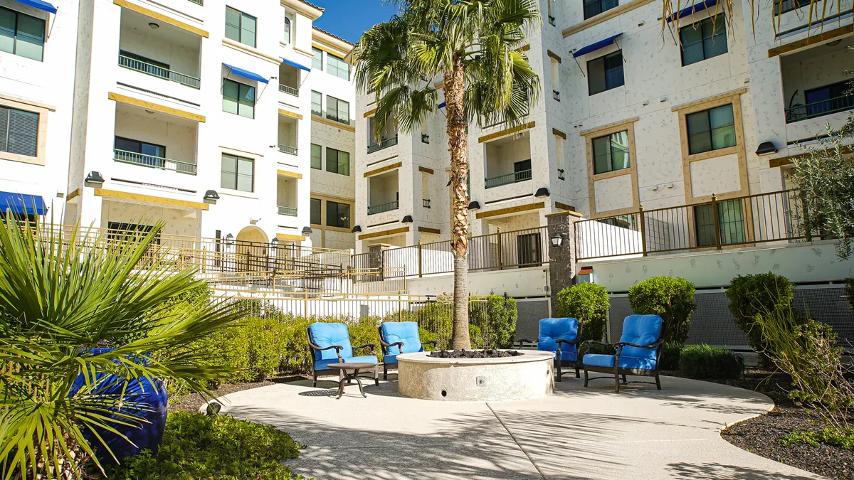 A relaxed communal outdoor area featuring a fire pit surrounded by blue chairs, lush greenery, and a central palm tree. The setting suggests evening gatherings, friendly conversation, and a welcoming environment for both residents and guests.