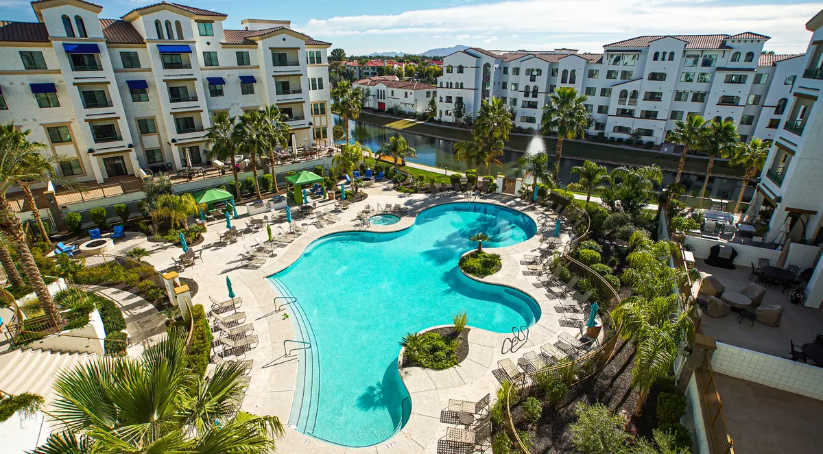 A vibrant resort-style swimming pool surrounded by palm trees, plush lounge chairs, and water features, with apartment residences visible in the background. The setting is lively with people swimming, sunbathing, and relaxing around the pool area.