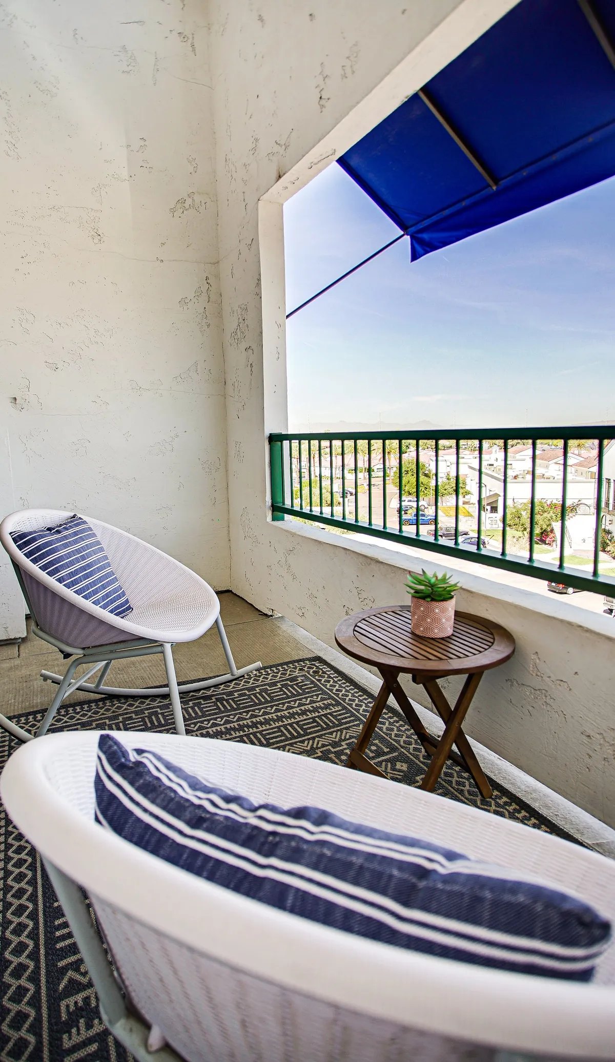 A cozy outdoor seating area features two white chairs with blue cushions, a small wooden table with a potted plant, and a blue awning overhead. The inviting patio space is perfect for enjoying a morning coffee, reading, or casual conversations, with a relaxed atmosphere ideal for families or groups to gather outside.