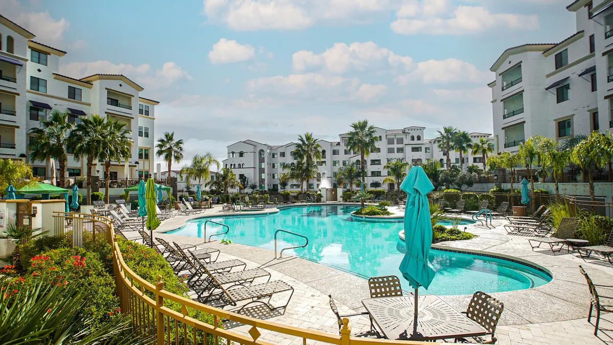 A stunning outdoor swimming pool encircled by tall palm trees, with inviting lounge chairs and green umbrellas set up poolside. The tranquil blue water reflects the sunny sky, while the surrounding greenery and palm trees create a resort-like ambiance perfect for relaxation and socializing.