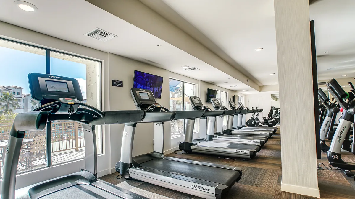 A bright and modern fitness center featuring rows of treadmills facing large windows, with flat-screen televisions providing entertainment. The room is filled with natural light, enhancing the motivational and energetic environment ideal for health and exercise routines.