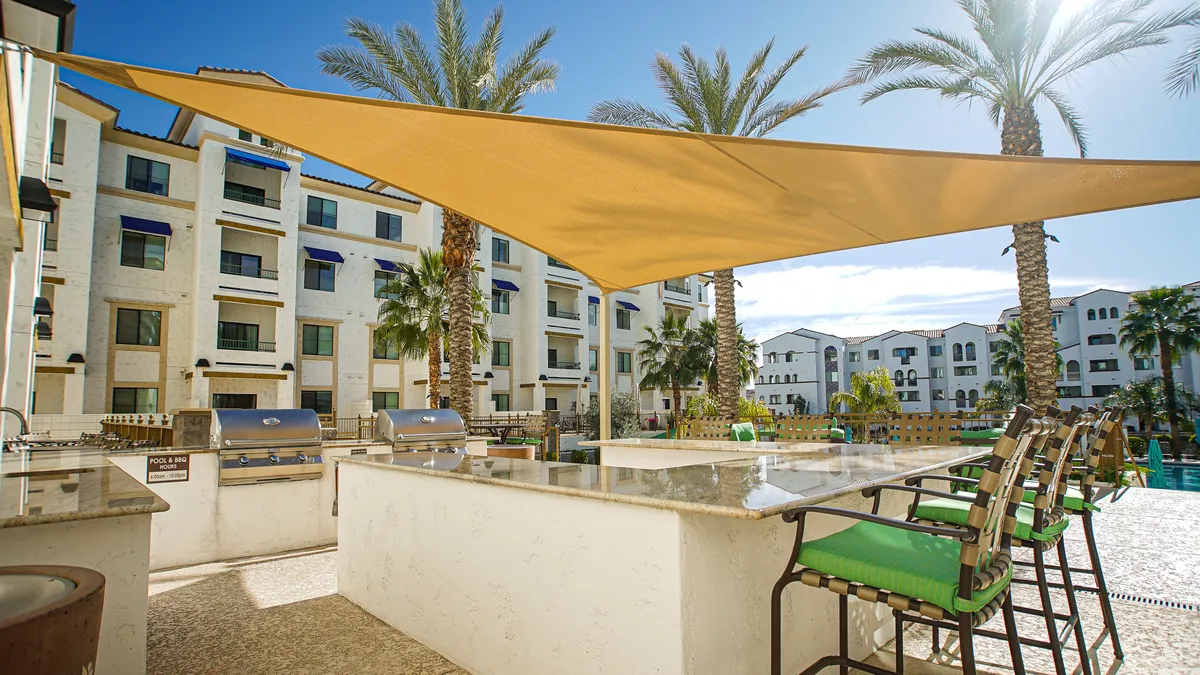 An outdoor grilling area adjacent to the pool, framed by palm trees and outdoor seating. People are seen barbecuing, socializing, sunbathing, and swimming nearby, creating a lively and communal ambiance.